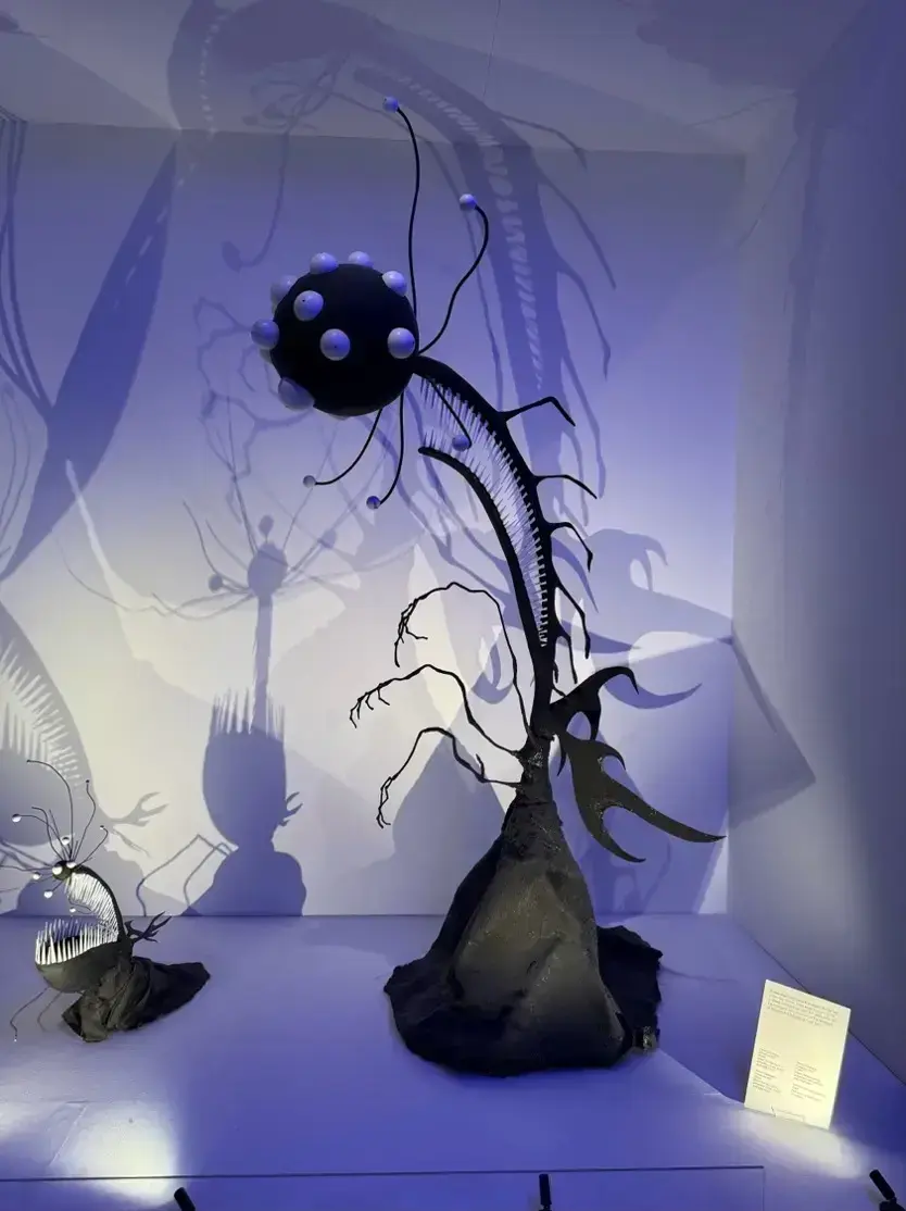 A dark sculptural exhibit featuring alien-like plant forms lit in deep blue, with dramatic shadows cast on the surrounding walls